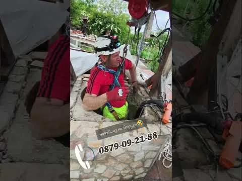 Майсторът със сбруя, каска и челник на ръба на каменен кладенец, над него виси телфера